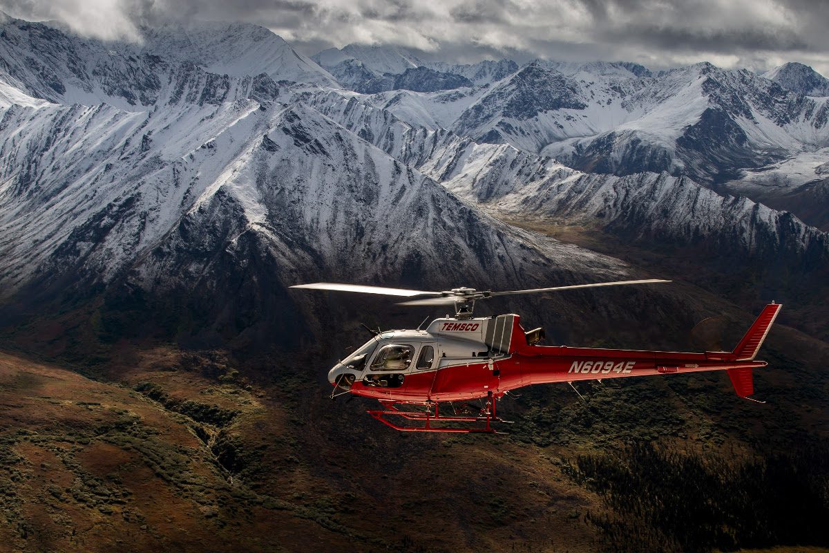 A helicopter flying over the mountains with snow on top.