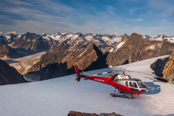Red helicopter on snowy mountain ridge with rocky peaks and glacier in background, under a blue sky.