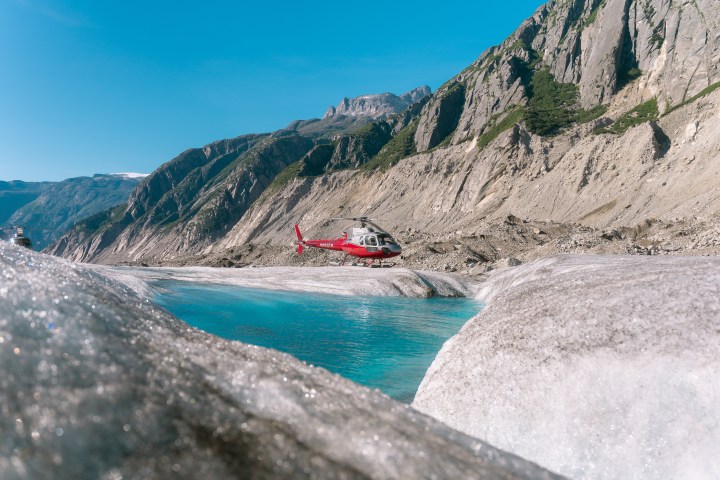 Red helicopter flying over a glacier with turquoise water and rocky mountains in the background.
