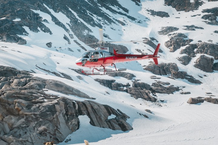 Red helicopter flying over snowy mountain with sled dogs in the foreground.