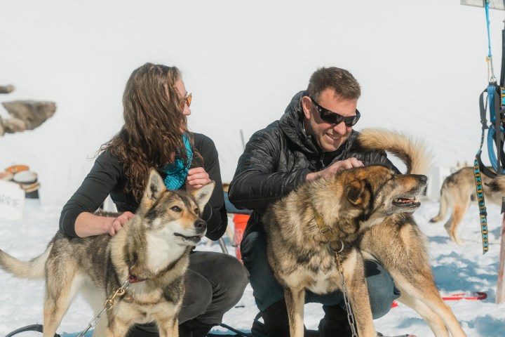 Two people sitting on snow, petting sled dogs with mountains in the background.