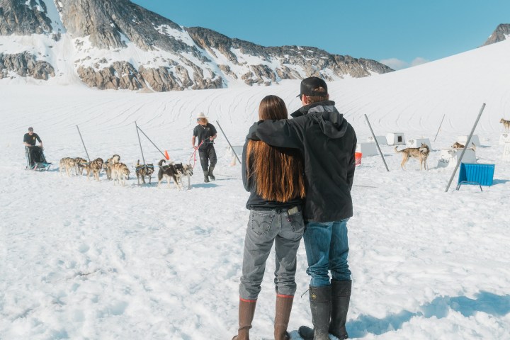 Couple on a snowy mountain watching sled dogs and mushers.