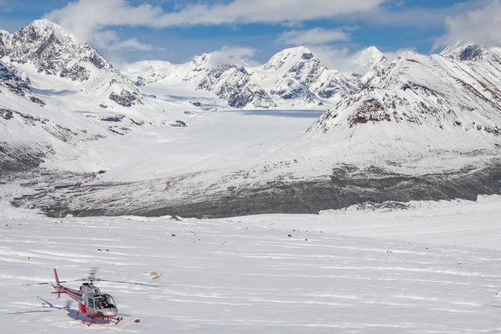 Red helicopter on snowy terrain with mountain range in the background under a clear blue sky.