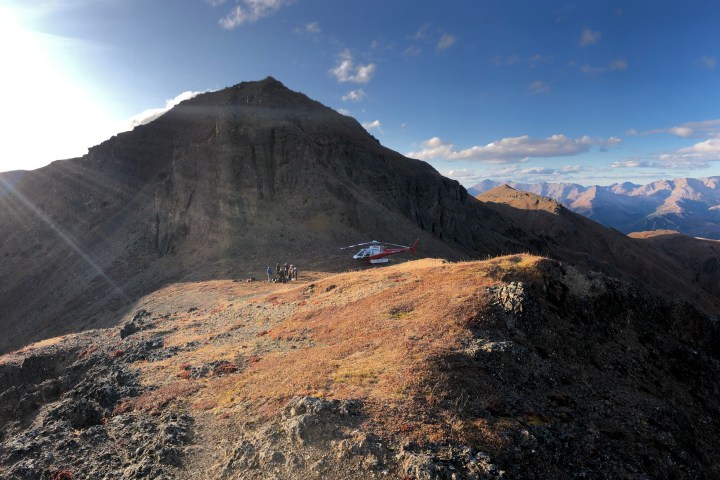 A helicopter on a mountain ridge during sunset, with people nearby and distant mountains in the background.