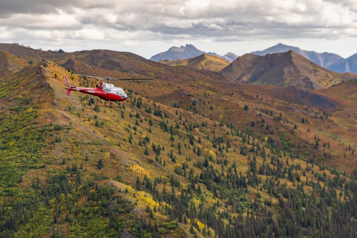 Red helicopter flying over colorful mountain landscape under cloudy sky.