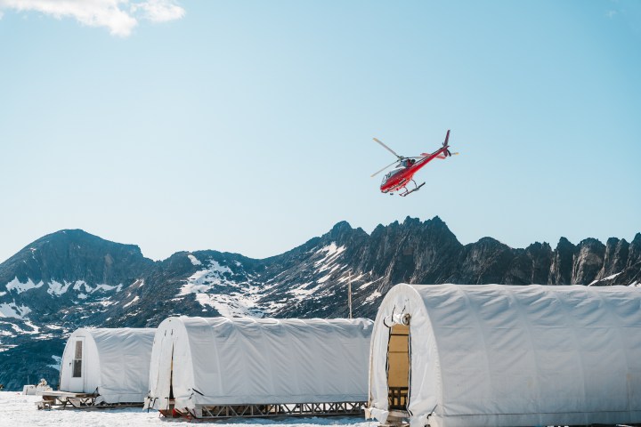 Red helicopter flying over snowy tents with rocky mountains in the background.