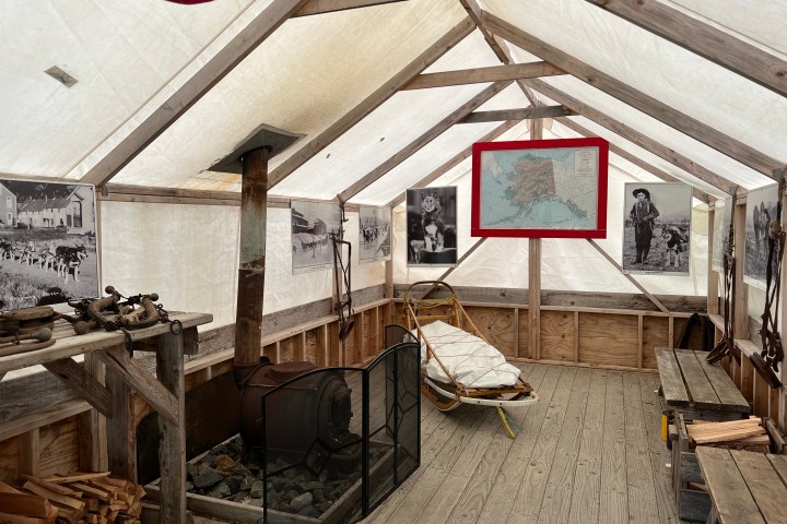 Inside view of a tent museum exhibit with photos, a sled, stove, and wood logs.