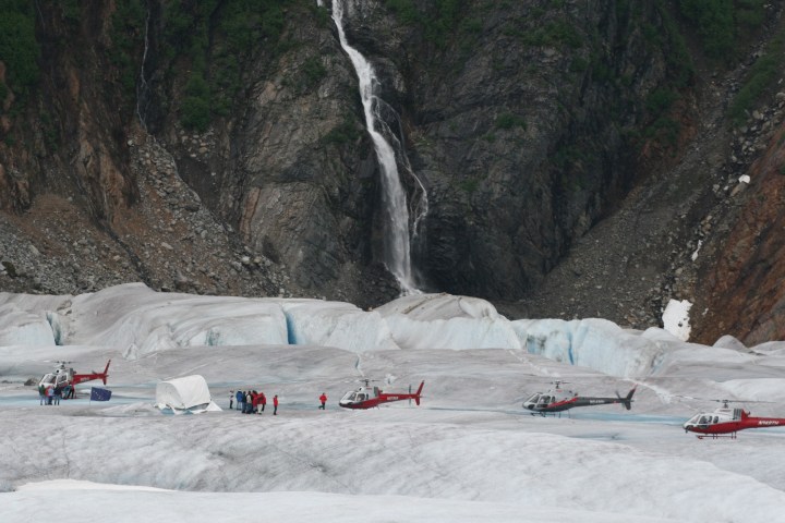 Helicopters on glacier with people near a waterfall at the base of a rocky cliff.