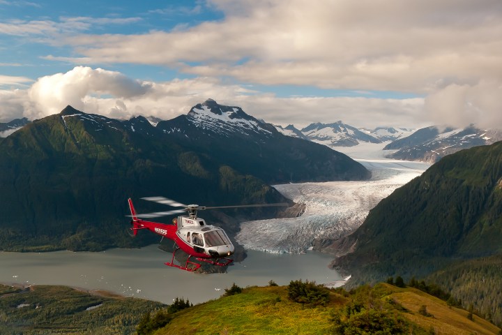 Red helicopter flying over a valley with mountains and a glacier in the background.