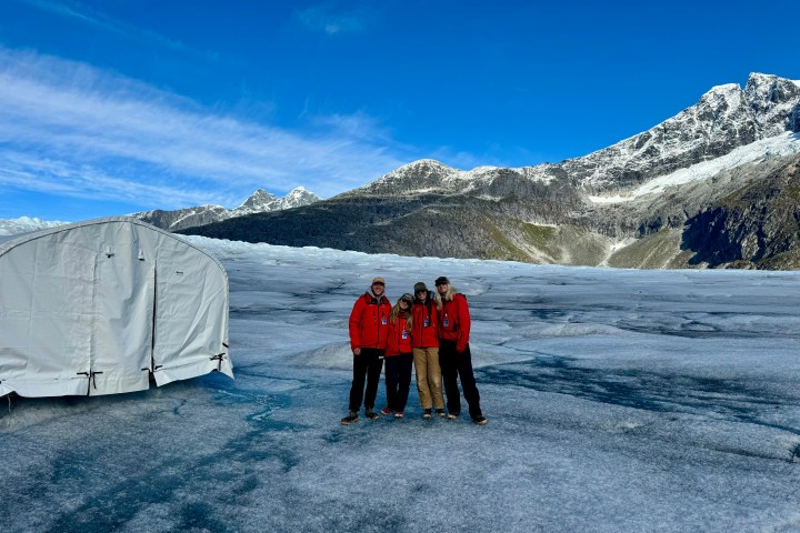 Four people in red jackets on a glacier with mountains and a white tent in the background.