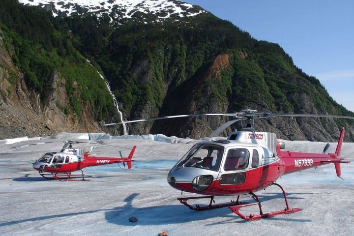 Two red helicopters on a snowy glacier with mountains and waterfall in background.