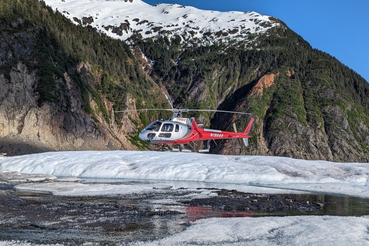 Red helicopter hovering over snow and ice in mountainous landscape.