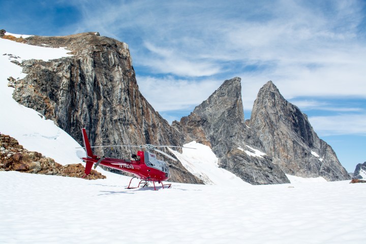 Red helicopter on snowy mountain terrain with rocky peaks under a blue sky.