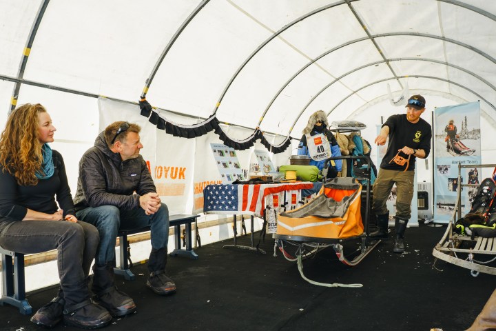 Three people in a tent with sleds and gear, one person gesturing while talking.