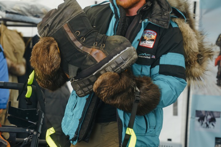 Person in fur-lined jacket holding a large boot inside a tent.
