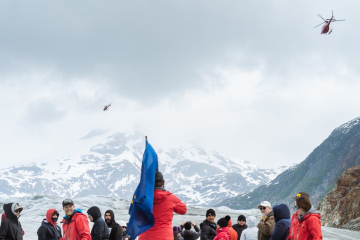 Group of people with blue flag on snowy mountain, two red helicopters in cloudy sky.