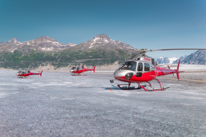 Three red helicopters on a snow-covered landscape with mountains in the background.