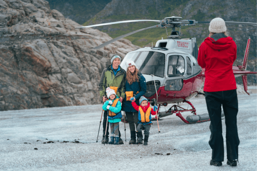 Family poses in front of helicopter on snowy terrain while person in red jacket takes a photo.