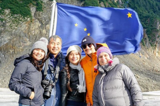 Group of five people smiling in front of an Alaska flag with a mountain backdrop.