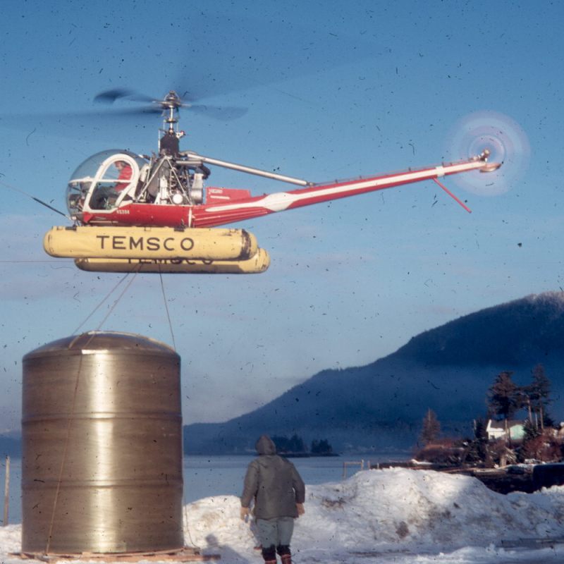 Helicopter lifting a large metal tank in a snowy landscape with mountains and trees.