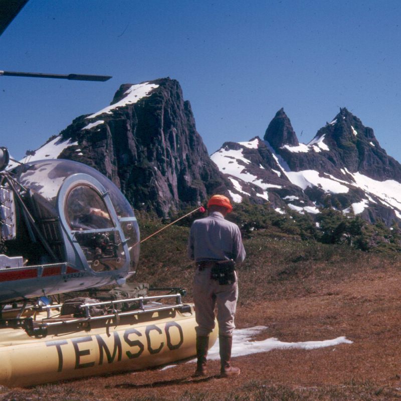 Helicopter and person near snowy mountain peaks under clear blue sky.