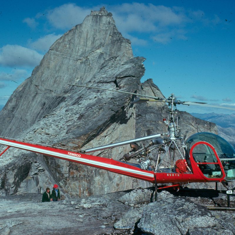 Red helicopter parked on rocky mountain peak with steep cliffs under a blue sky.